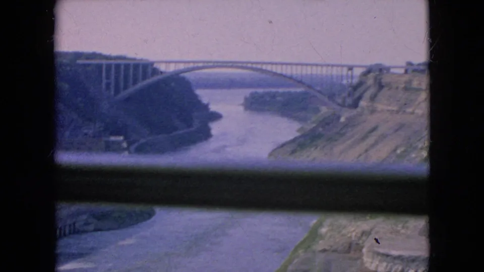 1964 Niagara Falls, New York footage frame with a large arched bridge crossing a broad river, with rocky slopes and wooded banks visible through a dark window opening.