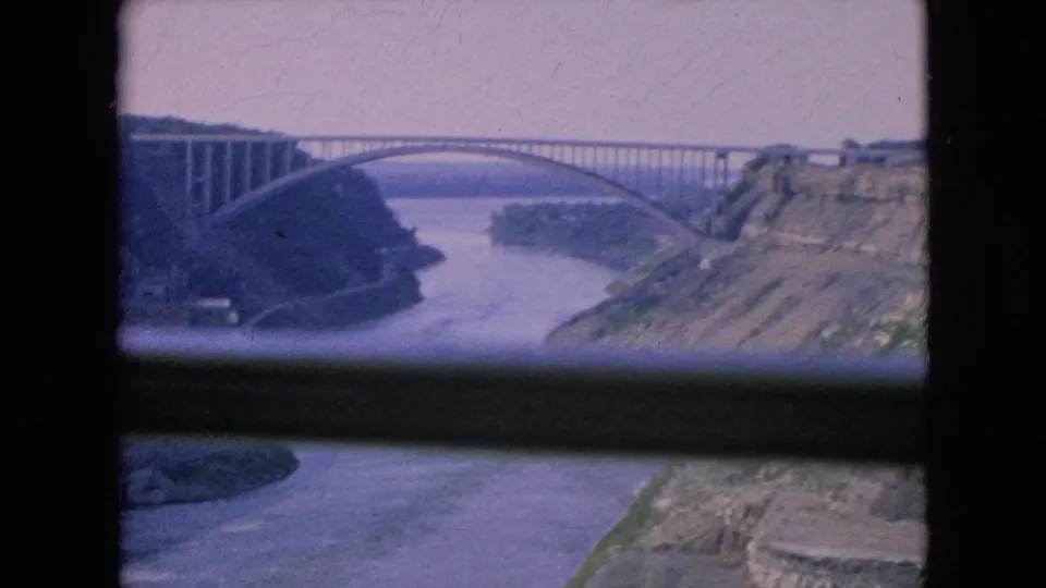 1964 Niagara Falls, New York home-movie frame showing the river gorge, an arched bridge, wooded banks, and dark foreground bars from the shooting vantage point.