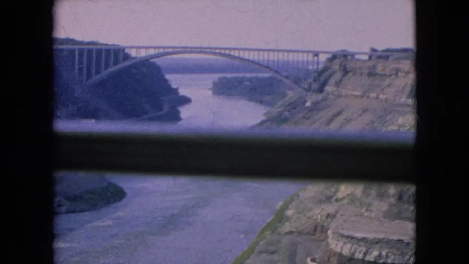 1964 Niagara Falls, New York frame showing a large arched bridge over a river gorge, with faded color, soft focus, rocky banks, and a dark foreground frame.