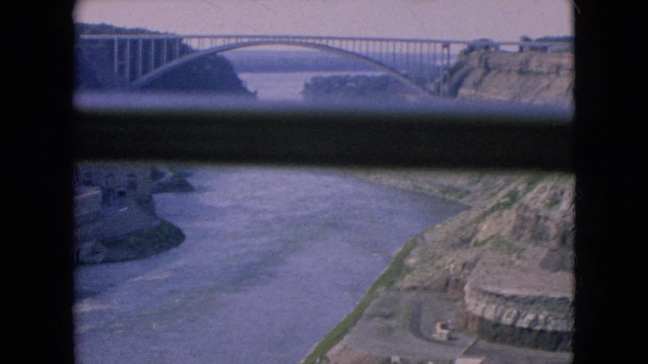 1964 8mm home-movie frame showing the Rainbow Bridge spanning the Niagara River, with gorge walls and dark window or railing bars across the view.