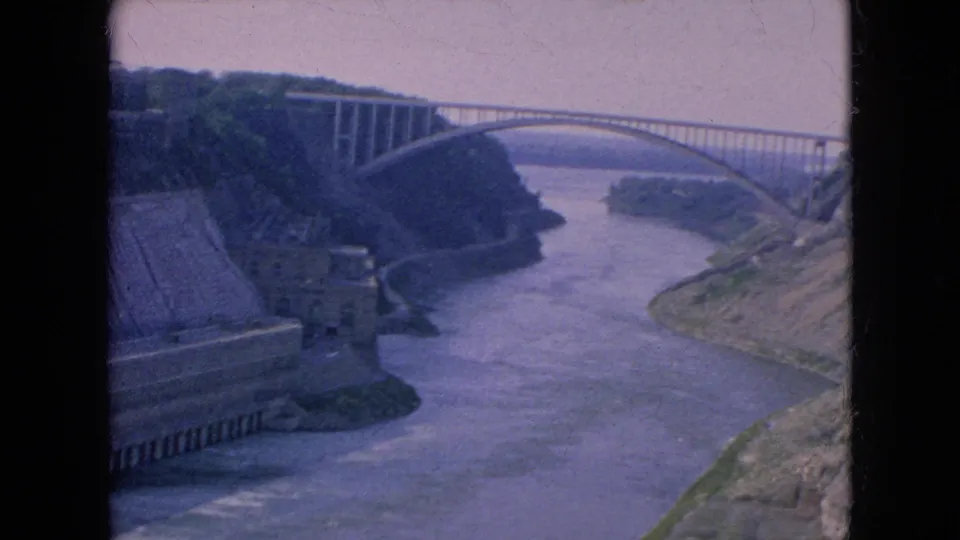1964 Niagara Falls home-movie frame with the Rainbow Bridge crossing the river gorge and concrete riverside buildings or walls visible on the left bank.