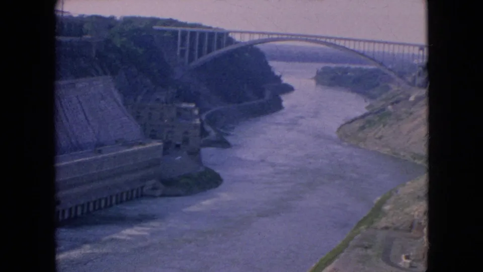 1964 8mm frame looking along the Niagara River toward the Rainbow Bridge, with a large concrete riverside structure, steep banks, and a roadway edge visible.