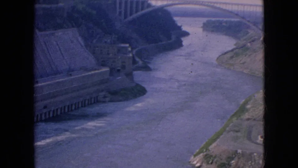 1964 Niagara Falls home-movie frame showing the Rainbow Bridge above the Niagara River, with the gorge, concrete riverside structure, and dark contact-sheet border visible.