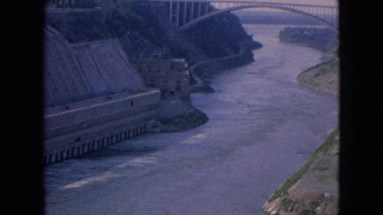 1964 8mm home-movie frame from the Niagara Falls, New York area showing a high-angle view of a concrete dam, river channel, and distant arched bridge.