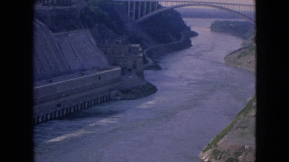 1964 8mm home-movie frame from the Niagara Falls, New York area showing a high-angle view of a concrete dam, river channel, and distant arched bridge.