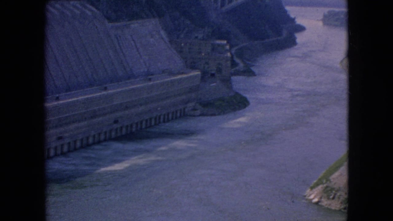 High-angle 1964 8mm view of the dam structure and controlled river channel in the Niagara Falls area, with an arched bridge visible beyond the waterway.
