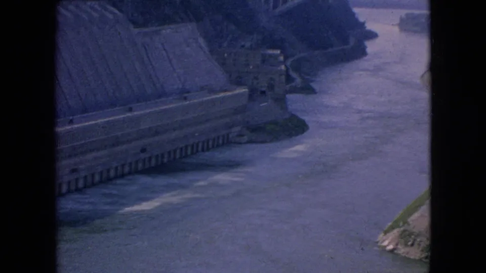 High-angle 1964 8mm view of the dam structure and controlled river channel in the Niagara Falls area, with an arched bridge visible beyond the waterway.