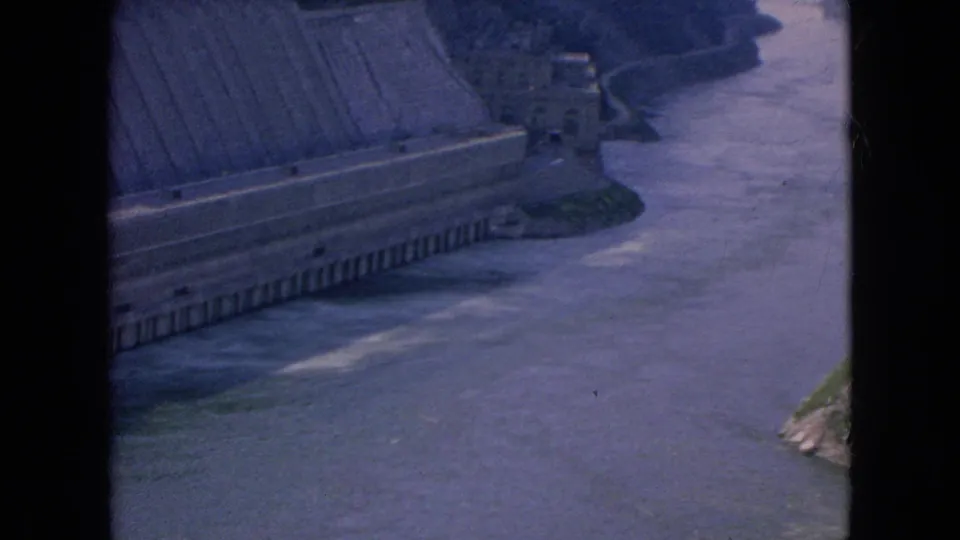 1964 8mm frame showing the dam edge, retaining structure, and flowing river below from an elevated visitor perspective in the Niagara Falls, New York area.
