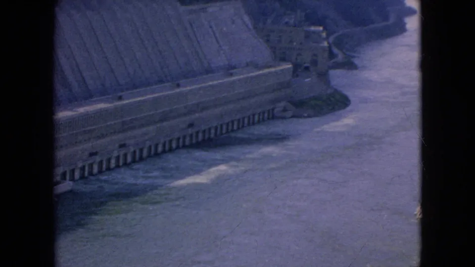 Final 1964 8mm frame looking over a concrete dam wall and curving river channel, with aged purple-blue film color and visible frame edges.