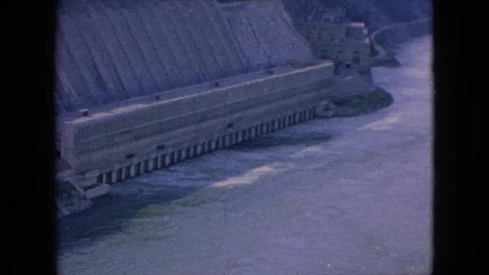 1964 8mm home-movie frame showing an elevated view over a broad river and a long concrete dam structure in the Niagara Falls, New York area.
