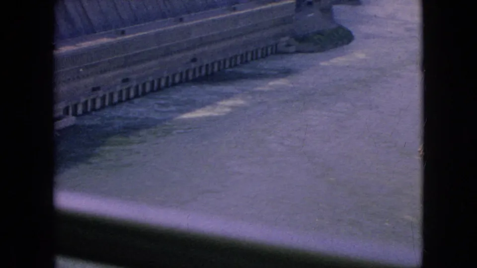 Angled visitor-shot frame of the concrete dam wall and water below, with faded blue-purple 8mm color and visible film-frame edges.