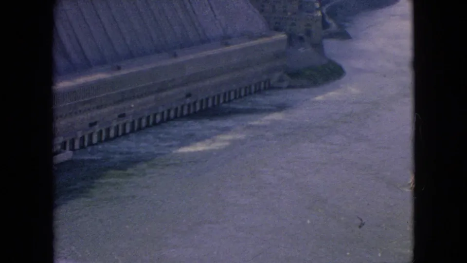 Final 1964 home-movie frame looking down on a long concrete dam beside the river, with the structure partly obscured by scan borders and soft 8mm focus.