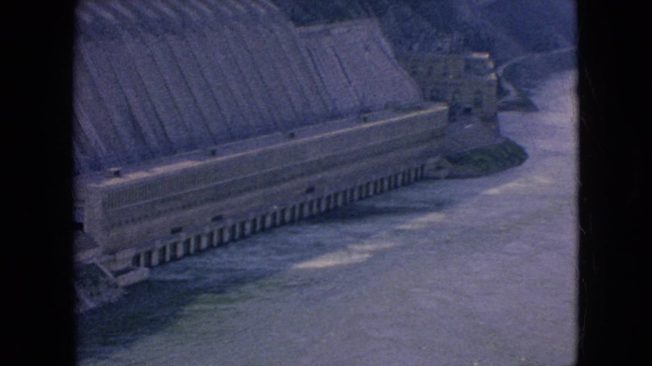1964 8mm view of a large concrete dam or power structure along the Niagara River area, with water and wooded slopes beyond.