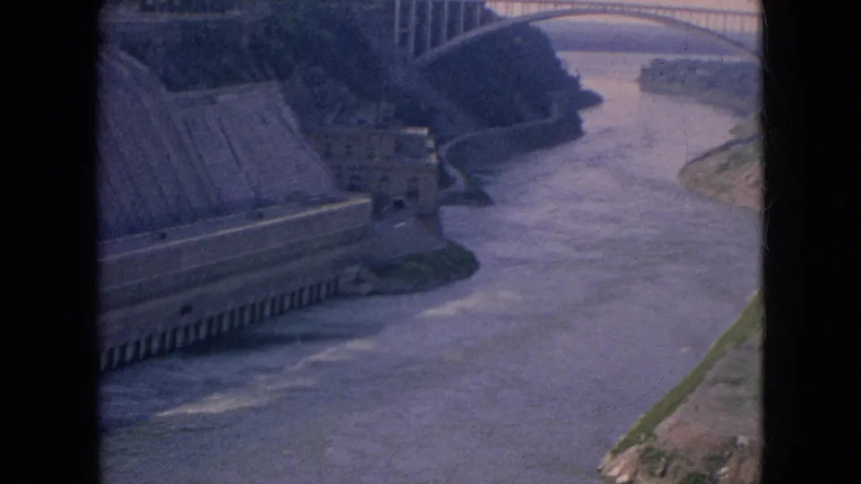 1964 8mm travel view looking past a concrete dam wall toward the river and an arched bridge across the valley.