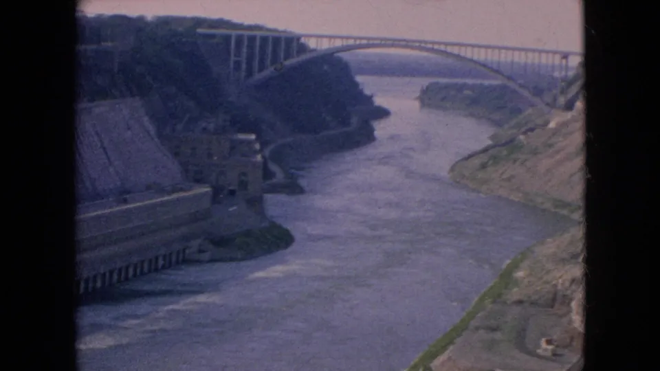 Wide 1964 home-movie view of the Niagara Falls, New York river valley, with a concrete infrastructure structure at left and an arched bridge spanning the water.