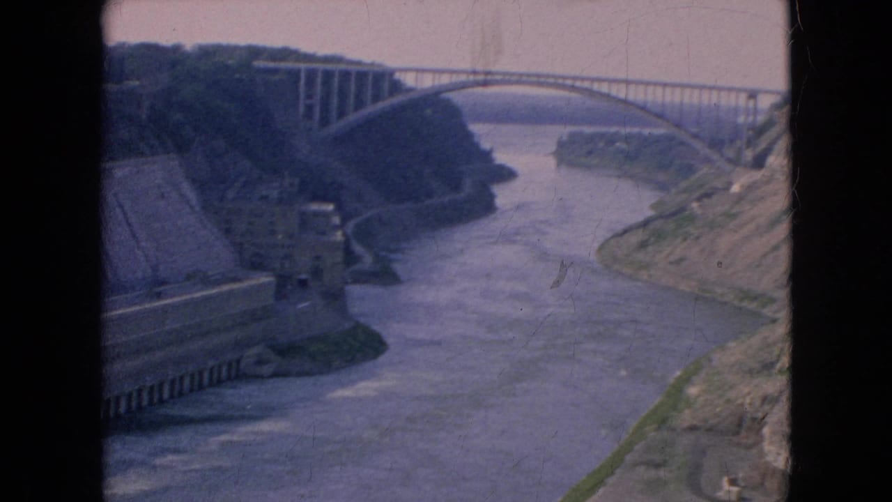 1964 8mm view of the Niagara River gorge near Niagara Falls, New York, with an arched bridge spanning the water and concrete infrastructure along the bank.