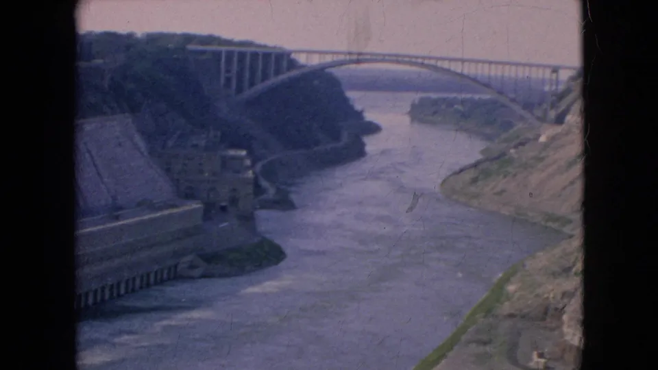 1964 8mm view of the Niagara River gorge near Niagara Falls, New York, with an arched bridge spanning the water and concrete infrastructure along the bank.