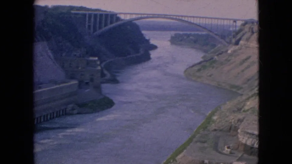 Wide 1964 home-movie frame of the Niagara River below a high arch bridge, with steep rock cuts and concrete works visible on both sides of the gorge.