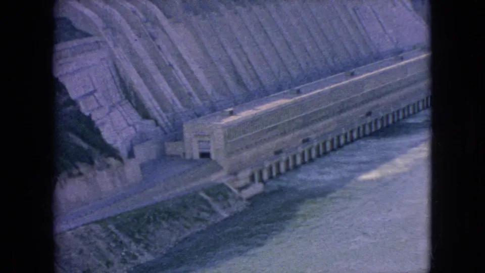 1964 8mm home-movie frame showing a large concrete hydroelectric dam at Niagara Falls, New York, with a stepped spillway and river water below.