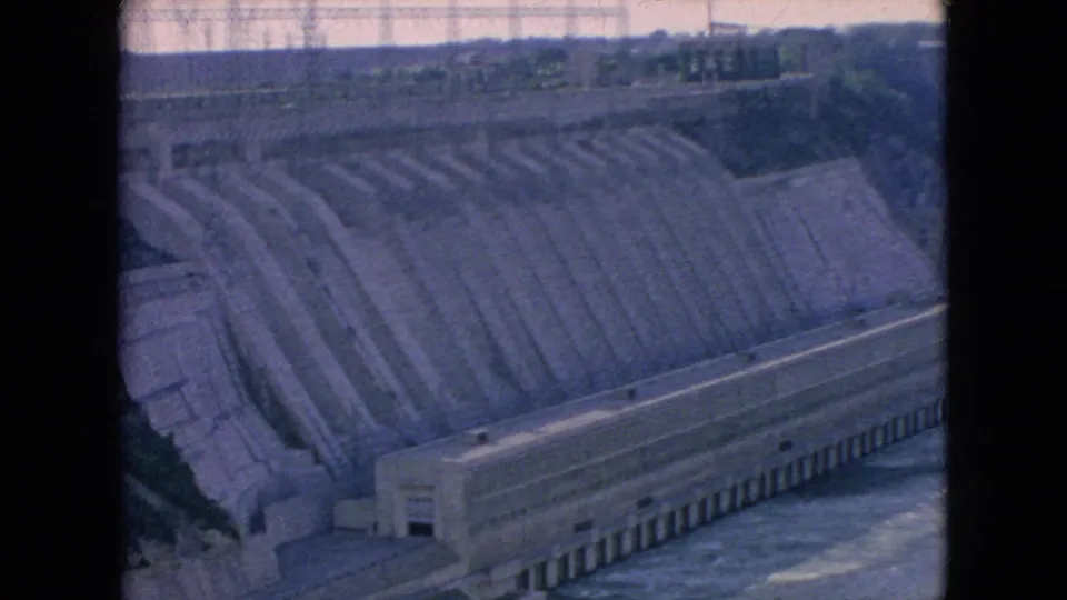 1964 8mm home-movie frame showing a wide view of the Niagara Falls hydroelectric dam, with stepped concrete surfaces, water below, and a distant tree line.