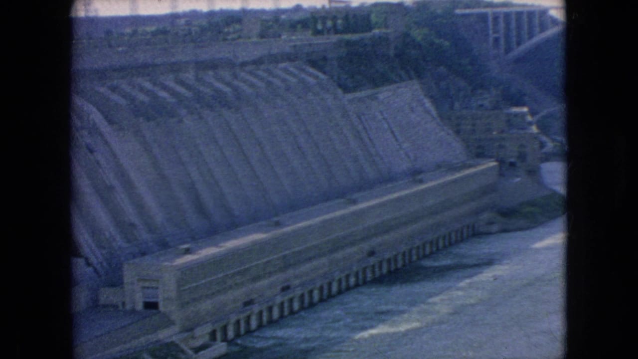 1964 Niagara Falls area home-movie frame with a large concrete spillway, base structure, river flow, and surrounding embankment visible.