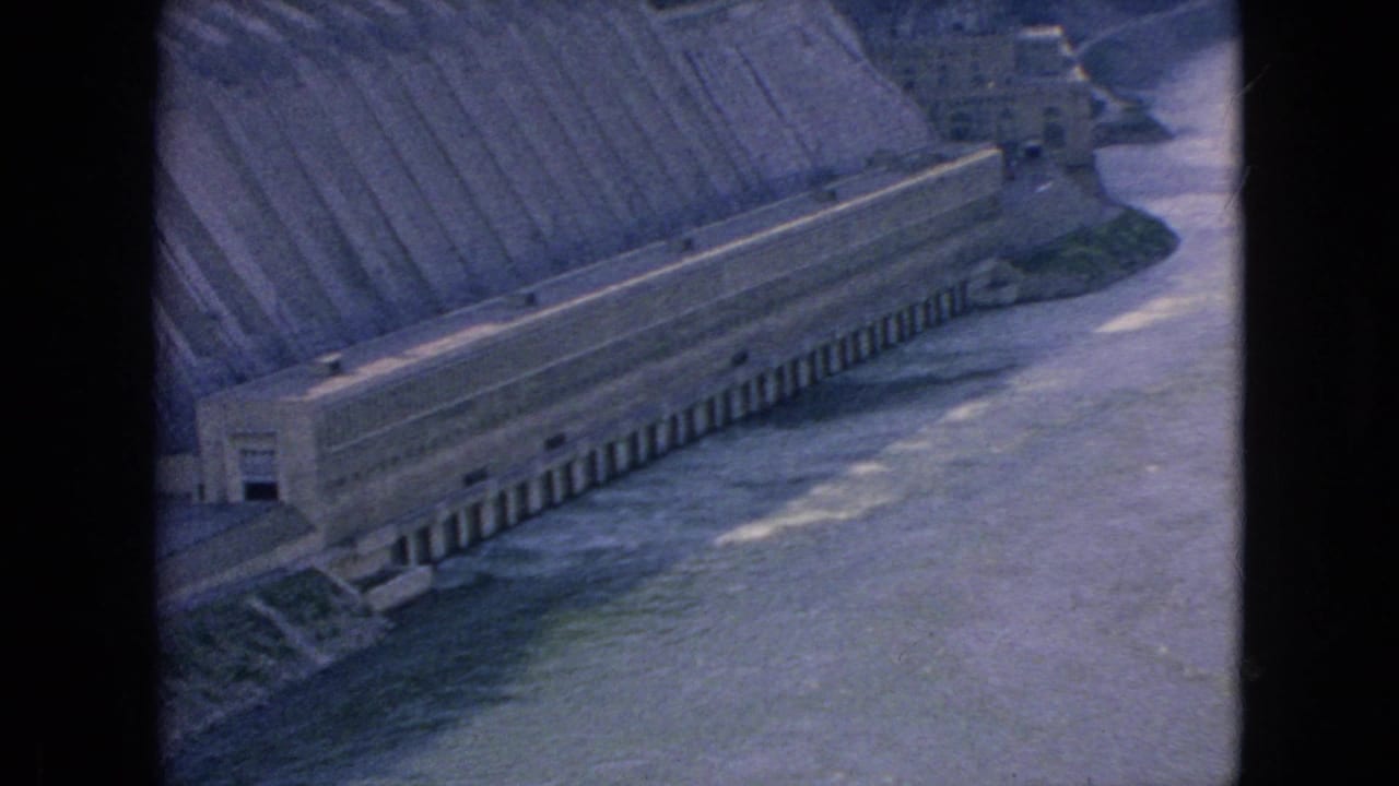 1964 8mm frame of the Niagara River area showing a large concrete dam structure, flowing water, and steep riverbanks from a high observation point.