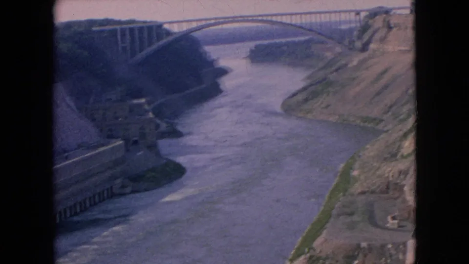 1964 Niagara River home-movie frame showing a long arched bridge in the distance, river water below, and dam infrastructure in the foreground.