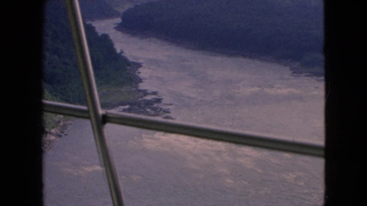 1964 8mm home-movie frame of a broad winding river below wooded bluffs, viewed through vertical and diagonal window-frame bars near Niagara Falls, New York.