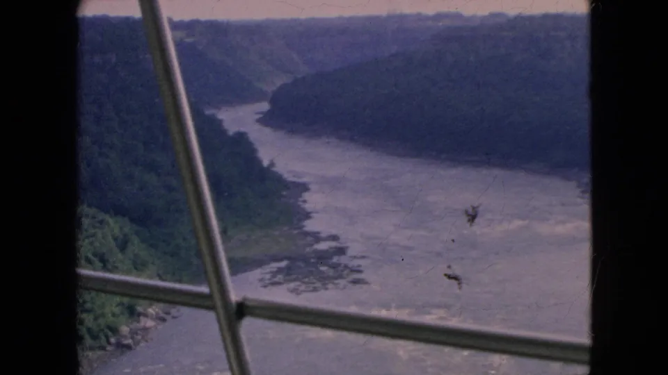 The river bend sits below steep green bluffs in this 1964 home-movie still, with visible film marks and window bars cutting across the scenic view.