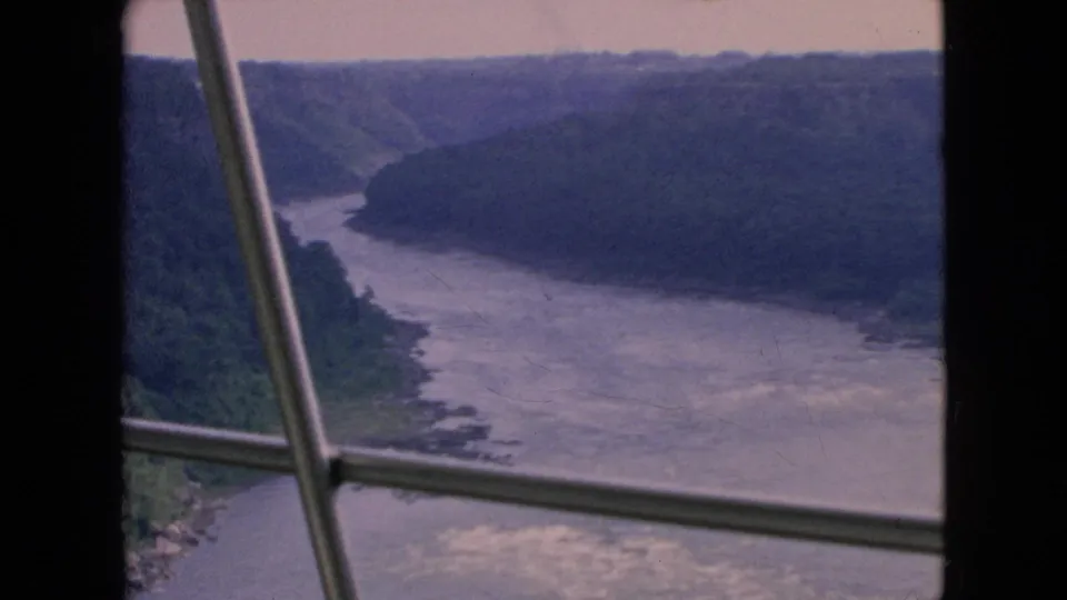 A steady 8mm frame shows a muted river valley and wooded cliffs under a pale sky, partially obscured by vertical and diagonal window-frame elements.