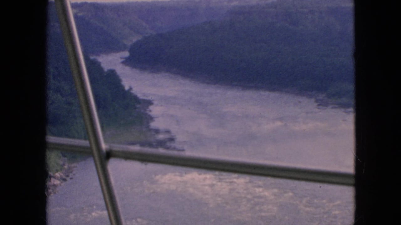 1964 8mm home-movie frame of a broad river with white water passing through a wooded gorge in the Niagara Falls, New York area, partially obscured by metal window or railing bars.