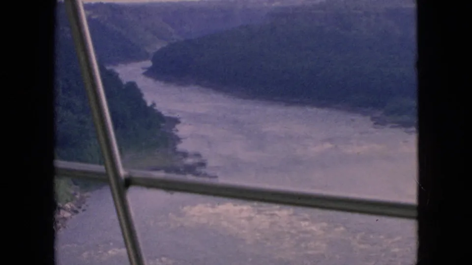 1964 8mm home-movie frame of a broad river with white water passing through a wooded gorge in the Niagara Falls, New York area, partially obscured by metal window or railing bars.