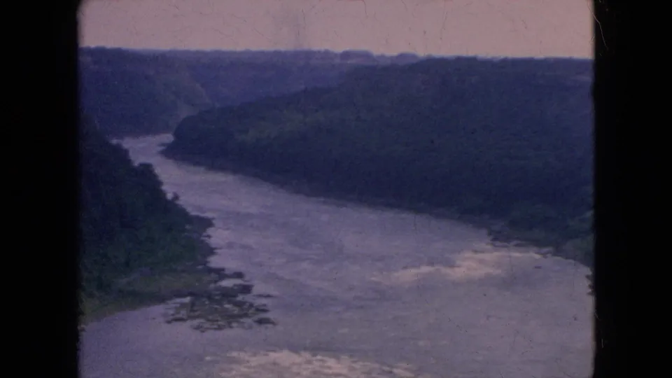Wide 1964 home-movie view of a river bend and white water below forested gorge walls, with the frame showing faded color and dark 8mm borders.