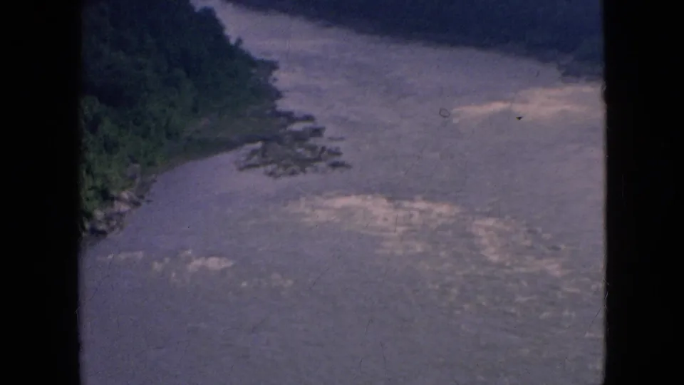 1964 aerial 8mm view of a forested riverbank, scattered rocks, and turbulent pale water along a broad river in the Niagara Falls, New York area.