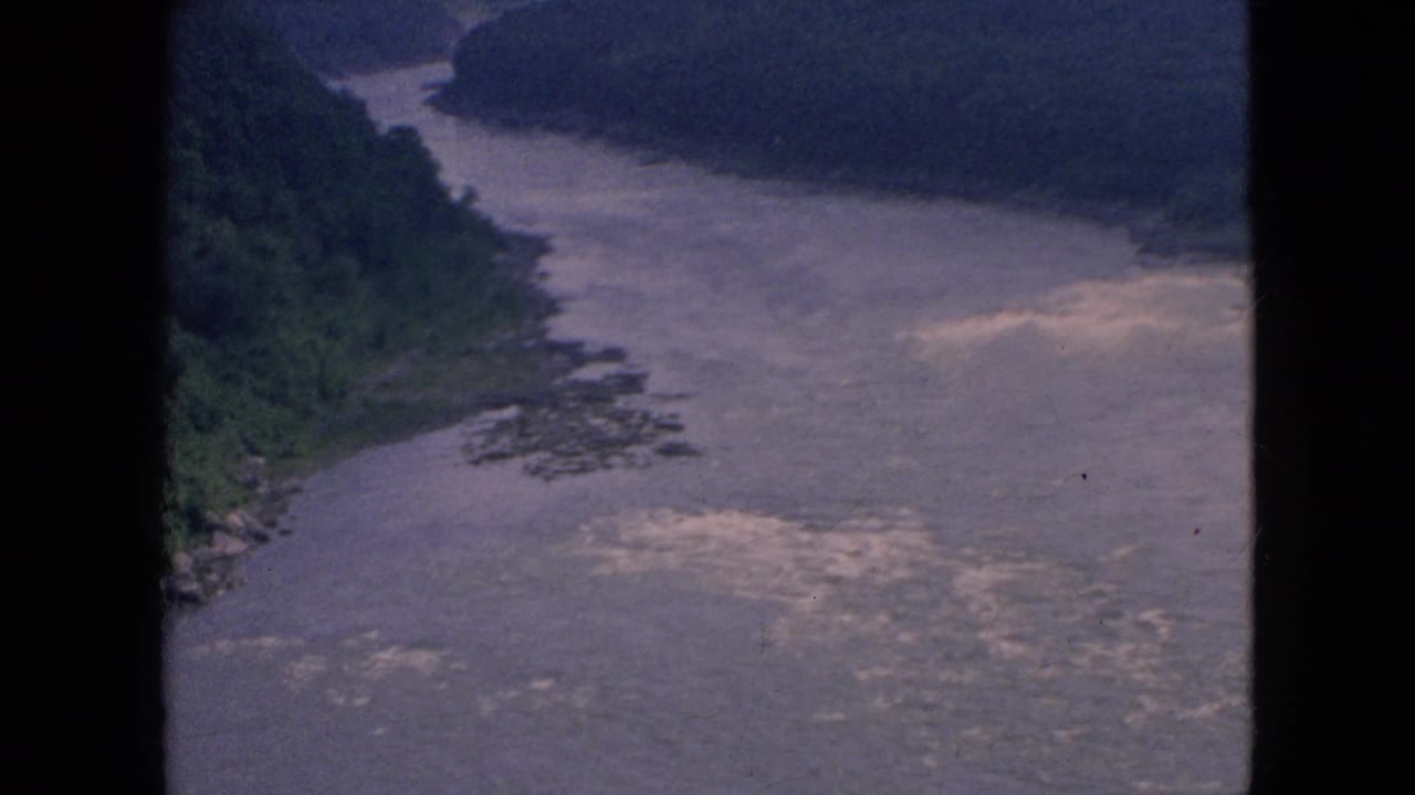 1964 home-movie frame of the Niagara River area from an elevated viewpoint, showing a broad water channel, wooded banks, and pale surface ripples.