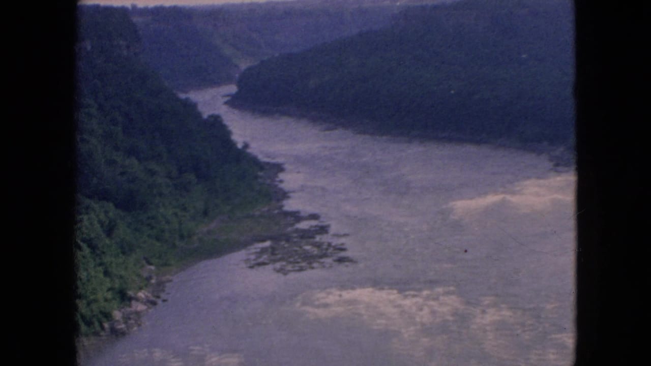 1964 Niagara-area 8mm frame showing the river curving through a wooded gorge, with muted color, soft focus, and bright patches on the water.