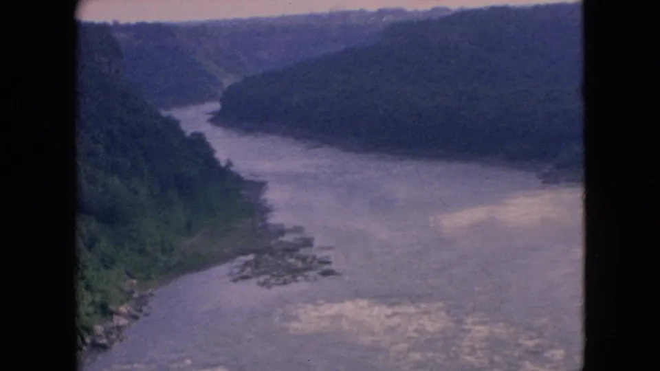 Soft 8mm frame of the Niagara River gorge in 1964, with green banks descending to a broad gray-blue river and faint rapids near the foreground.