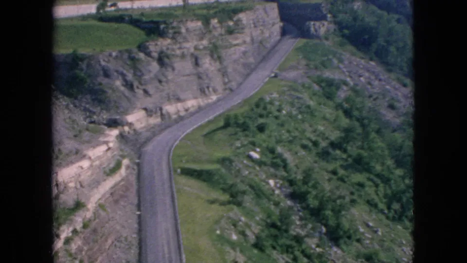 1964 8mm frame associated with Niagara Falls, New York metadata showing a curving road, rocky slope, green vegetation, and a dark vertical contact-sheet divider.