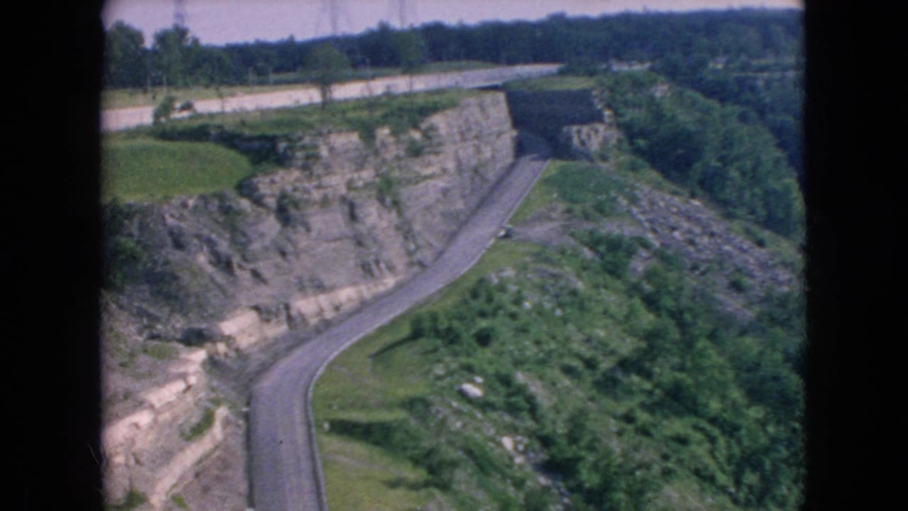 1964 8mm home-movie frame from the Niagara Falls, New York area showing a winding road cut below a rocky cliff with green slopes and trees.