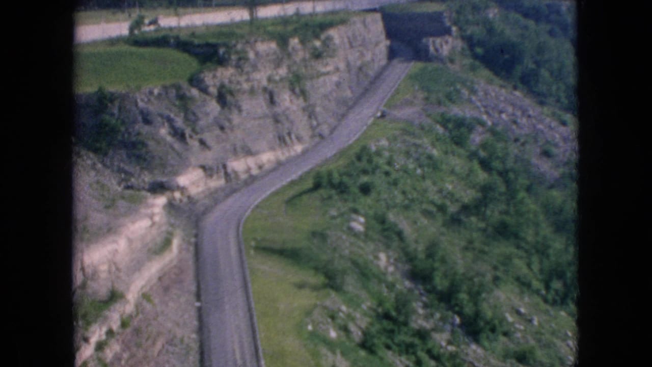 Curving road descends through a rocky slope with grass and trees around it.
