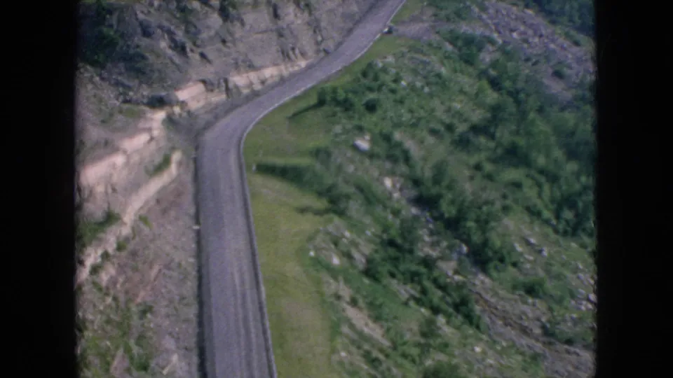 1964 8mm frame showing the cliffside road bending along a rock cut, with grassy ground and vegetation lining the roadway.