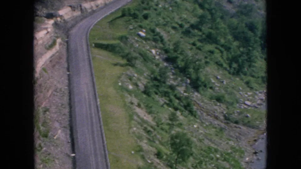 1964 Niagara Falls-area home-movie frame showing a road curving past rocky embankments, green grass, shrubs, and trees in the distance.