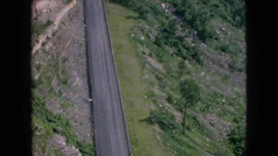 High-angle 8mm frame showing a narrow paved road alongside sloped rocky ground, trees, and green hillside vegetation, supported as 1964 Niagara Falls, New York footage by metadata.