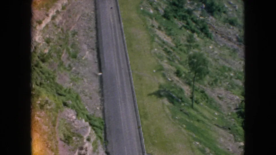 Vintage home-movie frame of a paved road descending through green hillside terrain with exposed rocks, roadside posts, and tree shadows in the supported 1964 travel context.