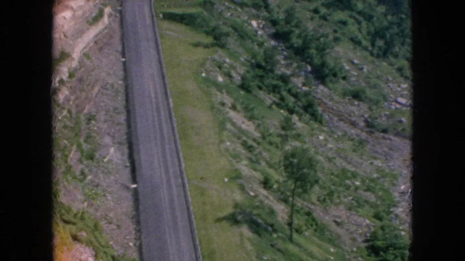 High-angle 8mm view of a paved road bordered by grass, rocky ground, and dense greenery, with dark film borders visible in the 1964 Niagara Falls, New York clip metadata context.