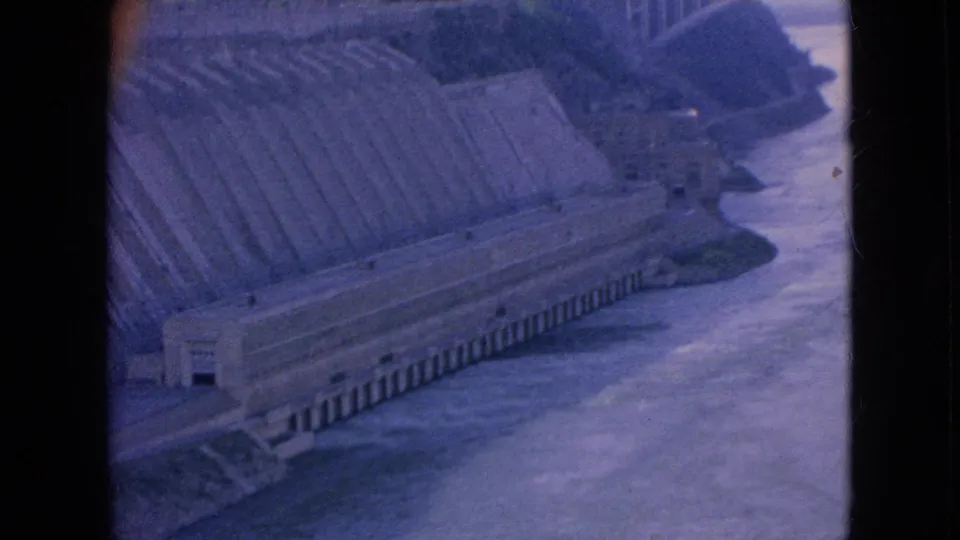 1964 Niagara Falls-area frame showing a large concrete dam or hydroelectric structure along the river, filmed from above with blue-purple 8mm color shift.