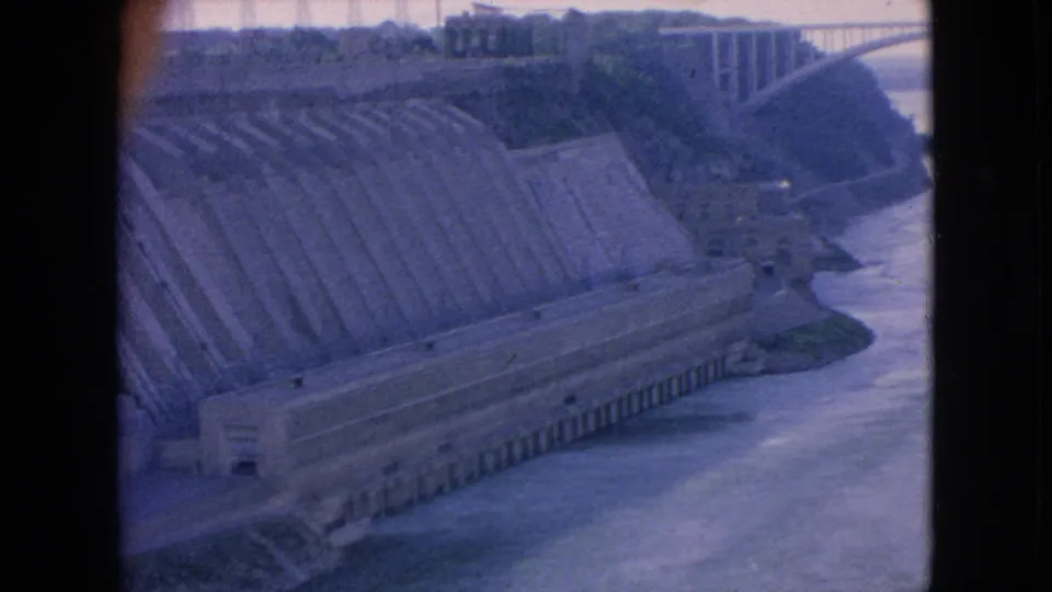 1964 Niagara Falls-area home-movie frame with a closer elevated view of concrete dam works, retaining walls, river water, and distant bridge structure.