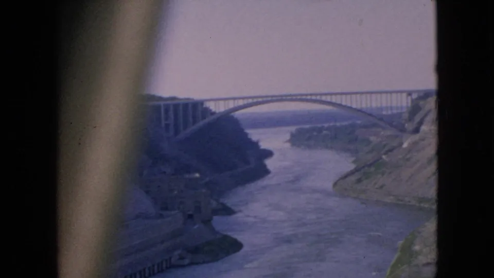 1964 Niagara Falls-area frame showing a long arched bridge spanning the river gorge above dam works and sloped embankments, with soft 8mm home-movie exposure.