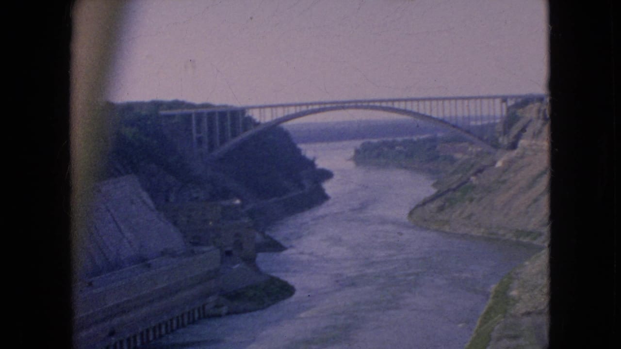 1964 8mm travel footage frame showing a high arched bridge over the Niagara River area, with steep banks and concrete works along the left shore.