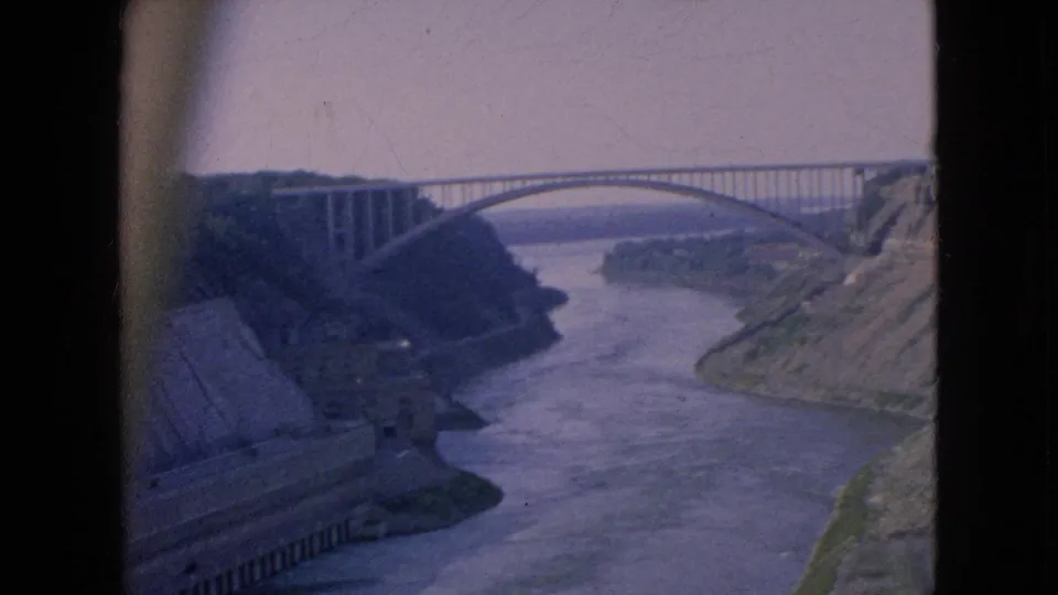 1964 8mm travel footage frame showing a high arched bridge over the Niagara River area, with steep banks and concrete works along the left shore.